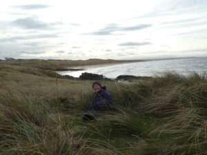 A woman sitting in long grass beside the sea.